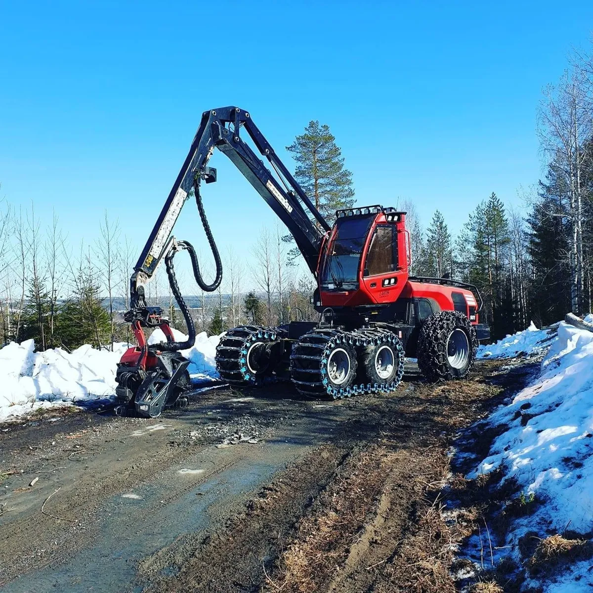 Röd skördare på en skogsbilväg mellan snövallar en solig vårvinterdag